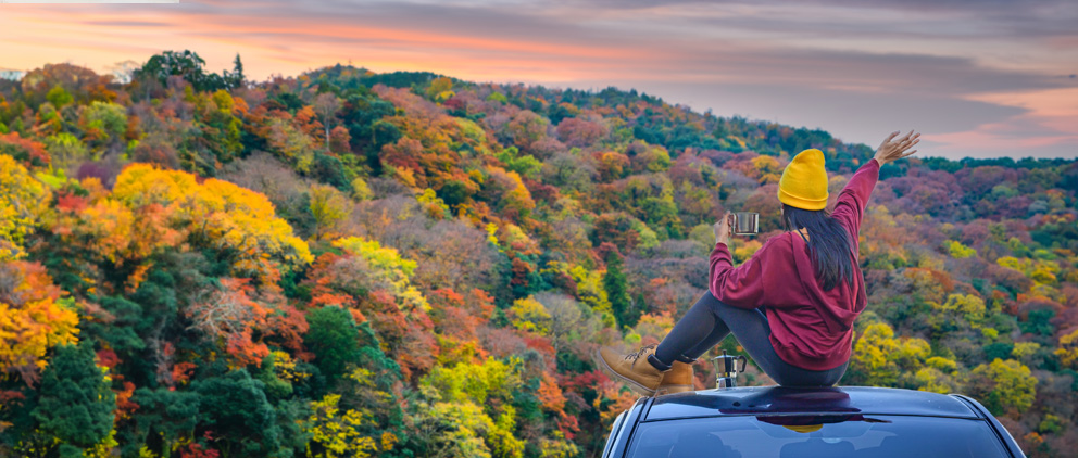 Woman sitting on the top of her car and enjoying a cup of coffee while enjoying a picturesque mountain vacation