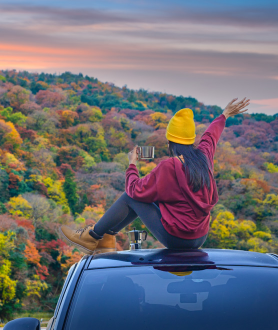 Woman sitting on the top of her car and enjoying a cup of coffee while enjoying a picturesque mountain vacation