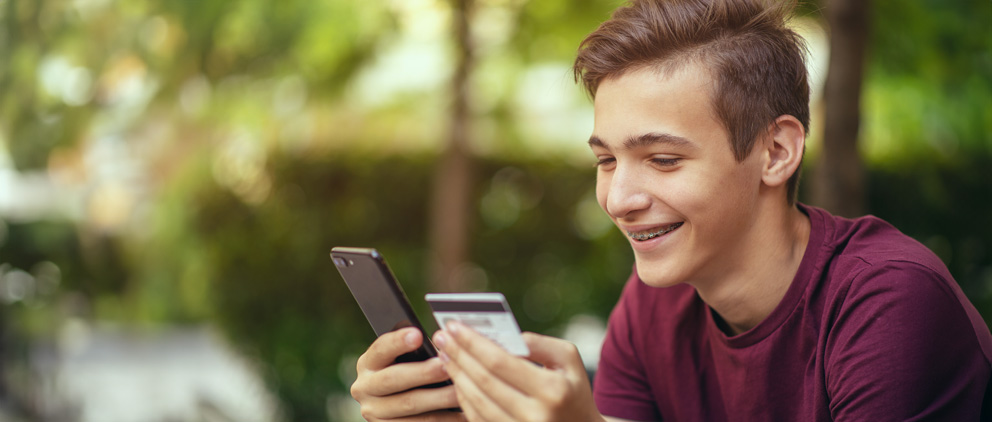 Teen boy holding his Visa Teen Debit Card and thinking about making a purchase with his phone