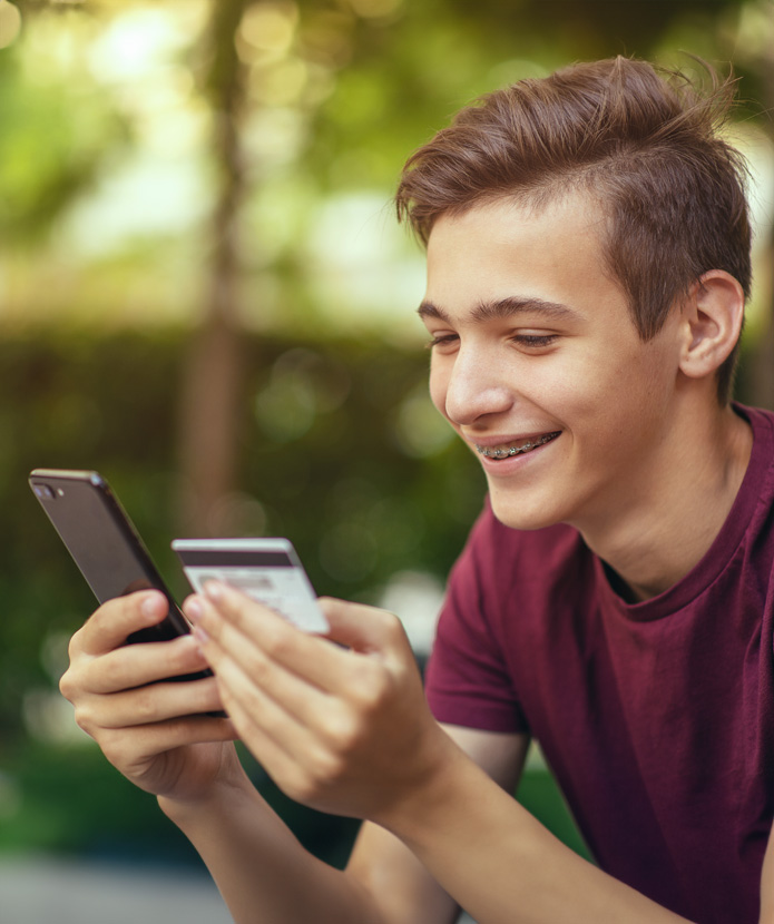 Teen boy holding his Visa Teen Debit Card and thinking about making a purchase with his phone